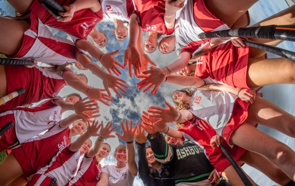 A team of young field hockey girls huddle up and put their hands in the middle for a team cheer on a cloudy day. 