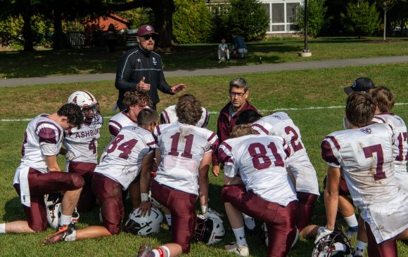 A team of Ashbury Football players in their uniforms all take a knee and surround the coach on the field.