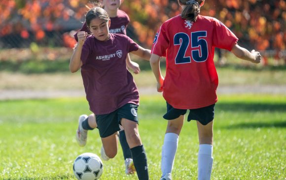 A young girl wearing an Ashbury top is kicking a soccer ball while another young girl defends on a bright autumn day. 