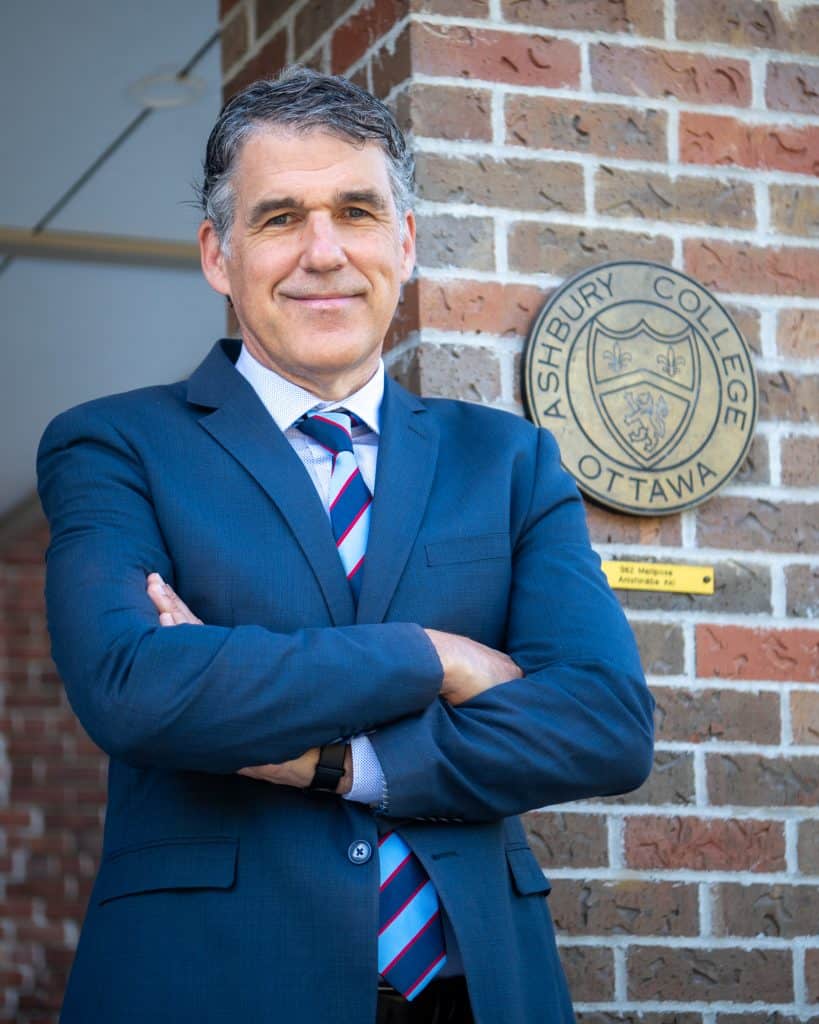 An older man wearing a blue suit is standing tall and proud, smiling with his arms crossed in front of a school. 
