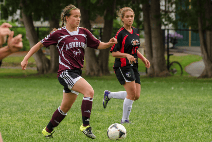 Emily Amano wearing an Ashbury soccer jersey is running up the field kicking a soccer ball with an opponent closely following her. 