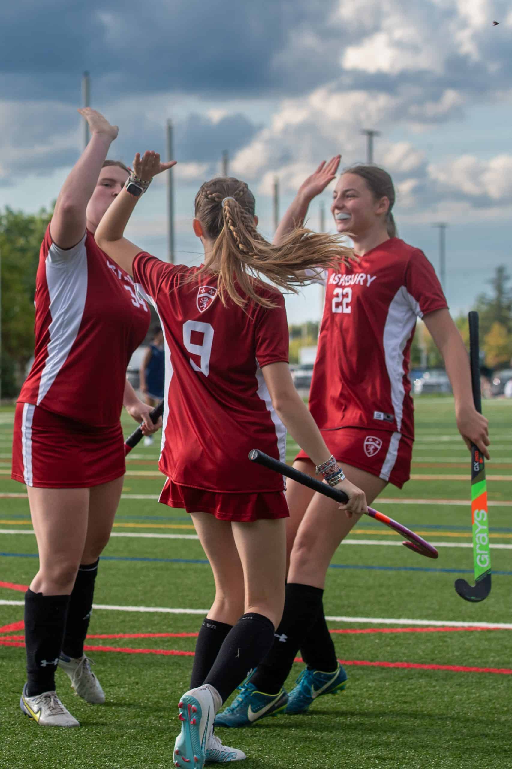 3 girls dressed in red Ashbury field hockey uniforms high-five and are smiling on a turf field